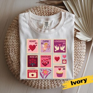 Op de afbeelding: Ivoor kleurig t-shirt met een verzameling postzegelontwerpen met Valentijnsdagthema. De postzegels bevatten harten, Cupido, tortelduifjes, rozen, chocolade en een cupcake. Het shirt ligt op een gevlochten mand.
