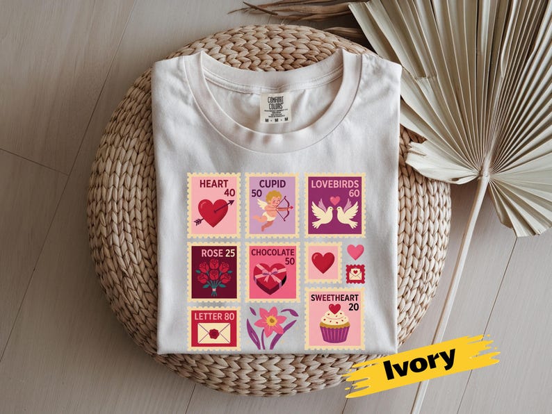 Op de afbeelding: Ivoor kleurig t-shirt met een verzameling postzegelontwerpen met Valentijnsdagthema. De postzegels bevatten harten, Cupido, tortelduifjes, rozen, chocolade en een cupcake. Het shirt ligt op een gevlochten mand.