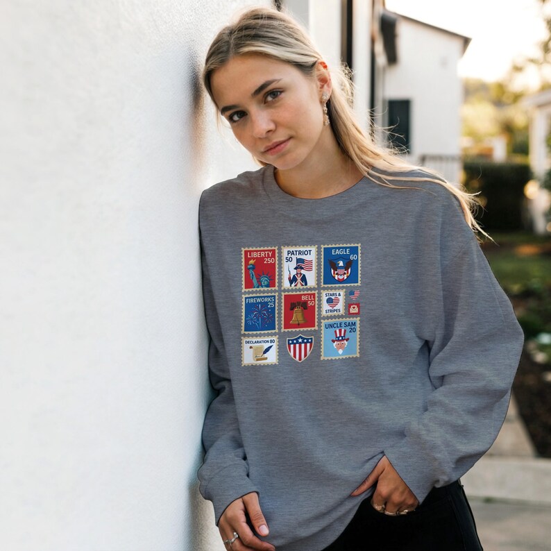 May include: A grey sweatshirt with a selection of colourful postage stamp designs. The stamps show patriotic symbols such as the Statue of Liberty, an American flag, and an eagle. The sweatshirt has a crew neck and long sleeves.