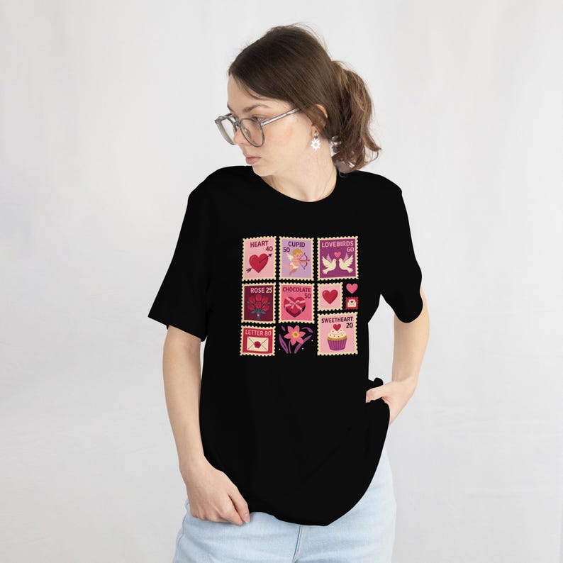 Op de afbeelding: Zwart T-shirt met een kleurrijk stempelontwerp met Valentijnsdagthema's. De postzegels tonen harten, Cupido, tortelduifjes, rozen, chocolade en een cupcake. Het shirt is gemaakt van een zacht materiaal.
