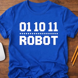 May include: A royal blue t-shirt with white pixelated text that reads "01 10 11" above "ROBOT". The shirt has short sleeves and a crew neck. A white computer mouse and two pencils are on a wooden surface.