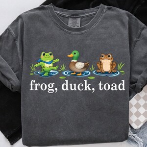 May include: Dark gray t-shirt featuring a whimsical design of a frog, duck, and toad in a pond, with the words "frog, duck, toad" printed below. The shirt has long sleeves and a crew neck.