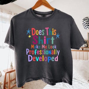 May include: A charcoal gray short-sleeve t-shirt featuring the multicolored text Does This Shirt Make Me Look Professionally Developed, accented with two blue striped stars and one small yellow star, printed on the front of the garment.