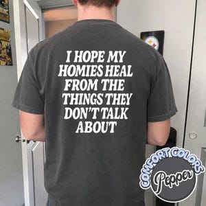 May include: A charcoal gray short-sleeve t-shirt featuring white text on the back that reads, I HOPE MY HOMIES HEAL FROM THE THINGS THEY DON'T TALK ABOUT. The shirt has a crew neckline and a relaxed fit, with a Comfort Colors Pepper label visible.