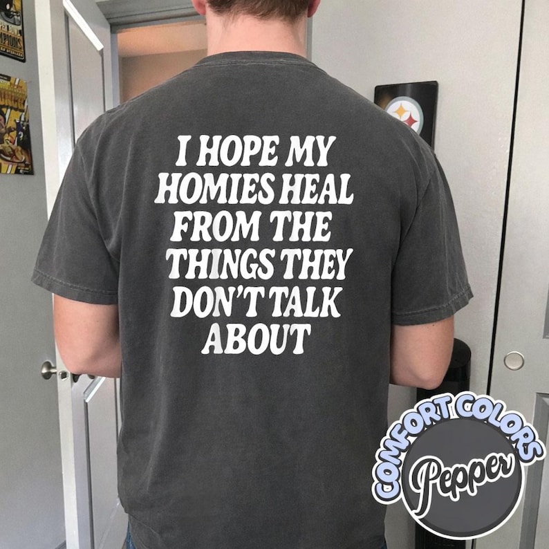 May include: A charcoal gray short-sleeve t-shirt featuring white text on the back that reads, I HOPE MY HOMIES HEAL FROM THE THINGS THEY DON'T TALK ABOUT. The shirt has a crew neckline and a relaxed fit, with a Comfort Colors Pepper label visible.