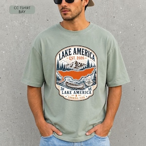 May include: A sage green short-sleeve t-shirt featuring a graphic print of a mountain landscape, a canoe, and a compass. The text reads LAKE AMERICA EST. 2026 LAKE AMERICA CANADA / USA. The design is enclosed in a decorative badge-style border.
