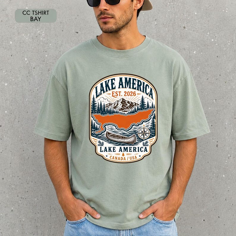 May include: A sage green short-sleeve t-shirt featuring a graphic print of a mountain landscape, a canoe, and a compass. The text reads LAKE AMERICA EST. 2026 LAKE AMERICA CANADA / USA. The design is enclosed in a decorative badge-style border.