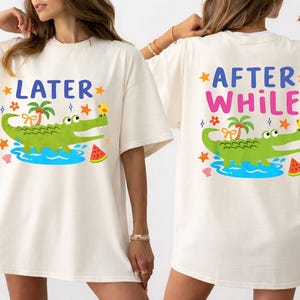 May include: Ivory-colored t-shirt featuring a cartoon alligator design. The front of the shirt has the word "LATER" above the alligator, while the back says "AFTER WHILE". The alligator is green, with a palm tree, a yellow bird, and watermelon slices.