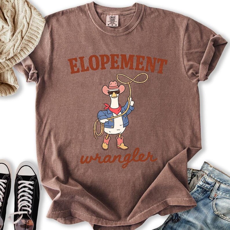 May include: Brown t-shirt with the word "ELOPEMENT" in orange letters. A cartoon goose wearing a cowboy hat, sunglasses, denim jacket, and boots is holding a lasso. The word "wrangler" is written below the goose.