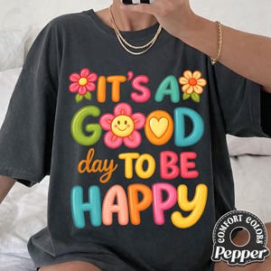 May include: Dark gray t-shirt with the phrase "IT'S A GOOD day TO BE HAPPY" in colorful, playful lettering. The design includes flower and heart accents. The shirt has a relaxed fit and a Comfort Colors brand tag.