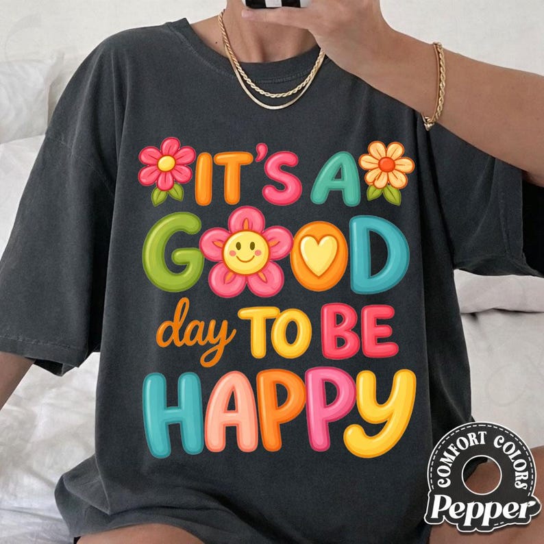 May include: Dark gray t-shirt with the phrase "IT'S A GOOD day TO BE HAPPY" in colorful, playful lettering. The design includes flower and heart accents. The shirt has a relaxed fit and a Comfort Colors brand tag.