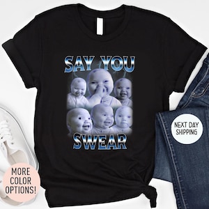 May include: Black t-shirt featuring a graphic of six babies with the text "SAY YOU SWEAR" in blue and white. The design has a lightning bolt effect. The shirt is paired with blue jeans and white sneakers.