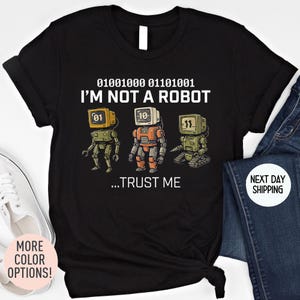 May include: Black t-shirt with a graphic of three retro robots with the text "I'M NOT A ROBOT ...TRUST ME" above the robots. The robots are in shades of green and orange with digital displays for heads. The shirt also has "MORE COLOR OPTIONS!" text.