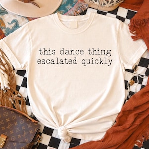 May include: Cream-colored t-shirt with the phrase "this dance thing escalated quickly" in black lettering. The shirt is knotted at the bottom. The background includes a black and white checkered pattern and a brown purse.