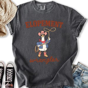 May include: Dark gray t-shirt featuring a cartoon goose wearing a cowboy hat, sunglasses, and a denim jacket, holding a lasso. The shirt has the words "ELOPEMENT" and "wrangler" in a rustic font.