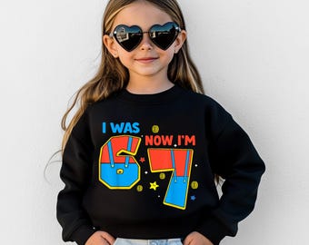 May include: A black sweatshirt with the text "I WAS 6 NOW I'M 7" in blue, red, and yellow. The number 6 and 7 are designed to look like overalls. The child is wearing heart-shaped sunglasses.