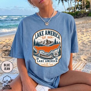 May include: A blue jean coloured t-shirt featuring a graphic print of a mountain landscape, a canoe, and a compass. The text reads Lake America, Est. 2026, and Canada/USA. The design includes pine trees and a central orange lake shape.