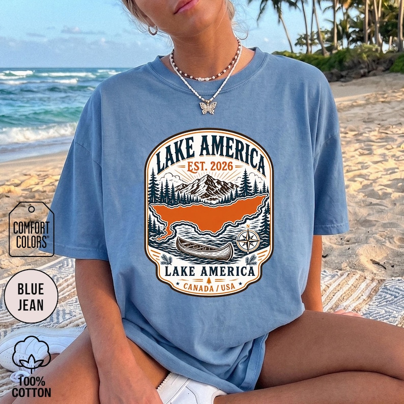 May include: A blue jean coloured t-shirt featuring a graphic print of a mountain landscape, a canoe, and a compass. The text reads Lake America, Est. 2026, and Canada/USA. The design includes pine trees and a central orange lake shape.