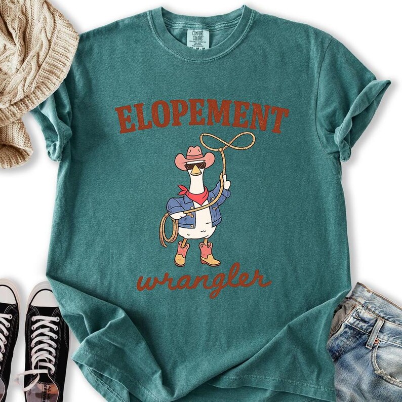 May include: Teal-colored t-shirt with the words "ELOPEMENT" and "wrangler" in a rust-colored font. The shirt features a cartoon goose wearing a cowboy hat, sunglasses, a red bandana, and a denim jacket, holding a lasso.