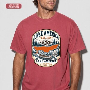 May include: A crimson short-sleeve t-shirt featuring a graphic print of a mountain landscape, a canoe, and a compass. The text reads LAKE AMERICA EST. 2026 and LAKE AMERICA CANADA / USA. The design includes pine trees, clouds, and a lake outline.
