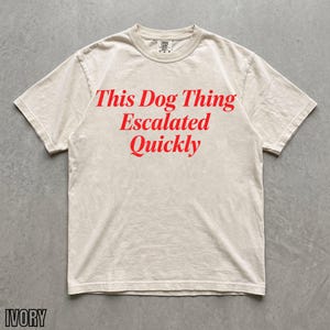 May include: An ivory short-sleeve t-shirt featuring the red text "This Dog Thing Escalated Quickly" printed in a serif font across the chest. The garment has a crew neckline and a standard fit, showcasing a clean and simple design for casual wear.