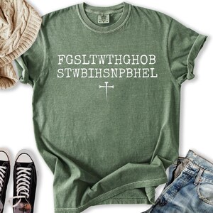 May include: A sage green t-shirt with white text and a cross graphic. The text is arranged in two lines with a cross below. The shirt is paired with blue jeans and black trainers.