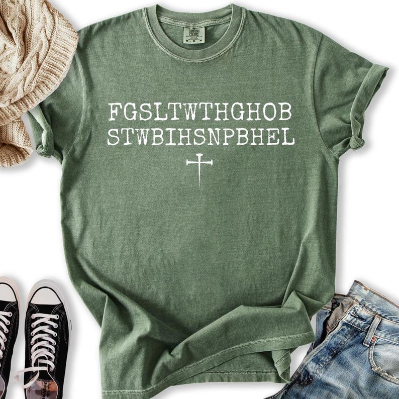 May include: A sage green t-shirt with white text and a cross graphic. The text is arranged in two lines with a cross below. The shirt is paired with blue jeans and black trainers.