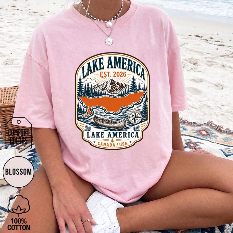 May include: A light pink t-shirt featuring a graphic print of a mountain landscape, a canoe, and a compass. The text reads LAKE AMERICA EST. 2026 CANADA / USA. The design includes pine trees and a large orange shape representing a lake body.