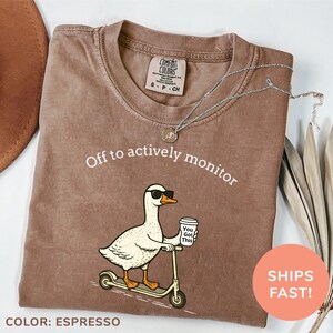 May include: Espresso-colored Comfort Colors t-shirt with the phrase "Off to actively monitor" and a cartoon goose on a scooter. The goose wears sunglasses and holds a coffee cup that says "You Got This."