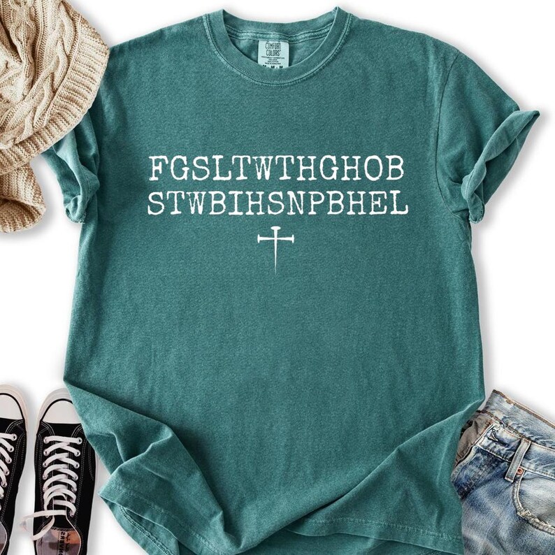 May include: Teal t-shirt featuring white text and a cross symbol. The text appears to be a coded message. The shirt is made of a soft material and has a crew neck. The shirt is folded, and a pair of black trainers are visible.