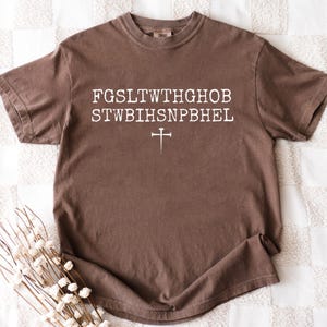 May include: A brown t-shirt featuring white text and a cross symbol. The text is arranged in two lines with a cross symbol below. The shirt is made of a soft material and has a classic crew neck design.