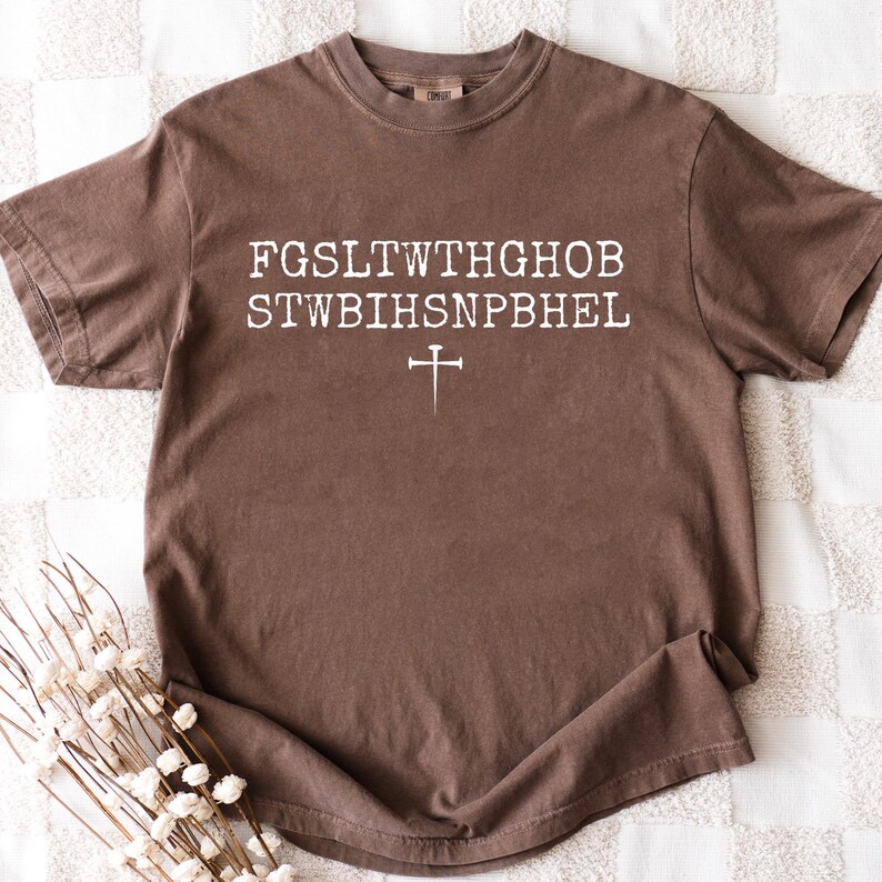 May include: A brown t-shirt featuring white text and a cross symbol. The text is arranged in two lines with a cross symbol below. The shirt is made of a soft material and has a classic crew neck design.