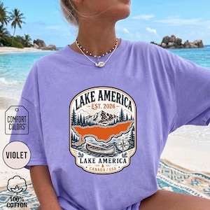 May include: A violet t-shirt featuring a graphic print with the text LAKE AMERICA EST. 2026 LAKE AMERICA CANADA / USA. The design includes a mountain range, a forest, a canoe, and a compass rose in shades of orange, dark green, and cream.