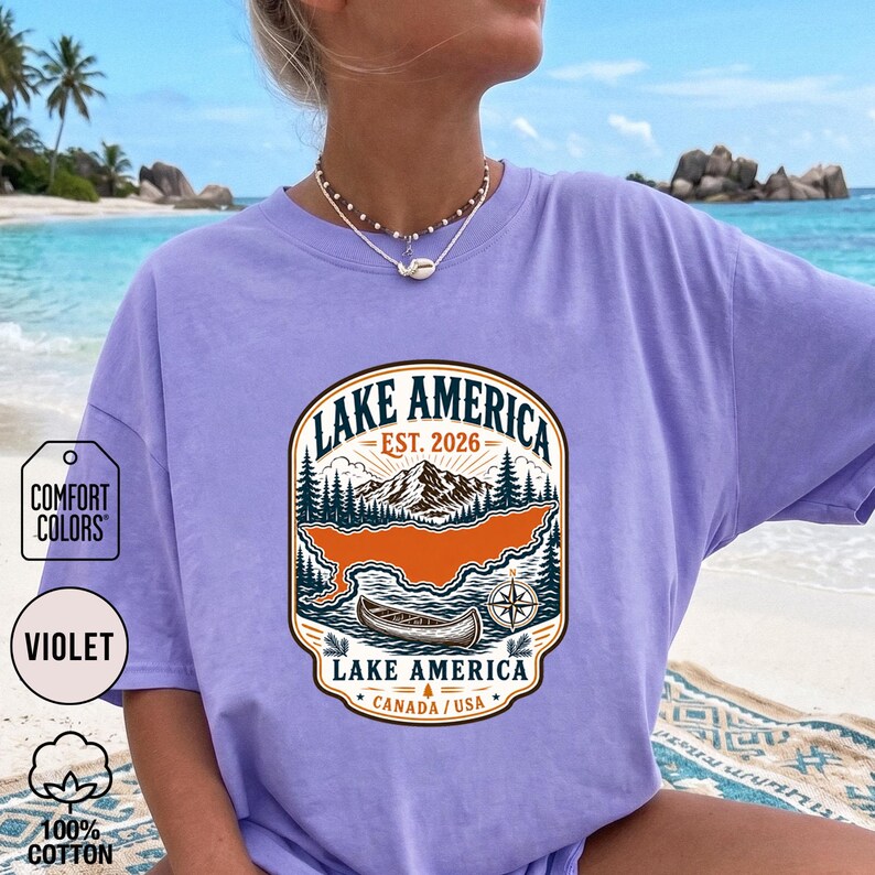 May include: A violet t-shirt featuring a graphic print with the text LAKE AMERICA EST. 2026 LAKE AMERICA CANADA / USA. The design includes a mountain range, a forest, a canoe, and a compass rose in shades of orange, dark green, and cream.