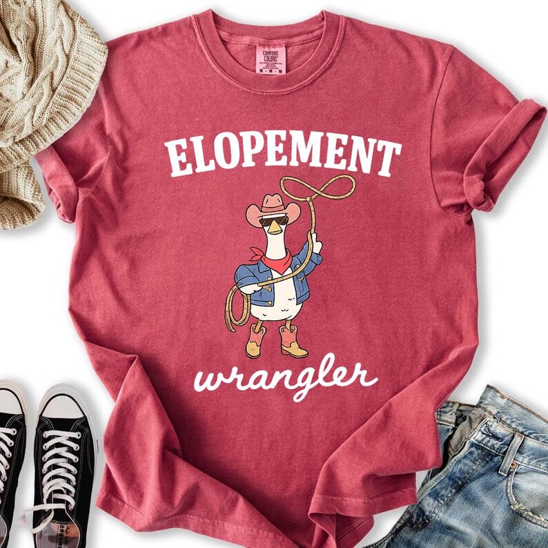 May include: A red t-shirt with the words "ELOPEMENT" and "wrangler" in white. The shirt features a cartoon goose wearing a cowboy hat, sunglasses, a denim jacket, and cowboy boots, holding a lasso.