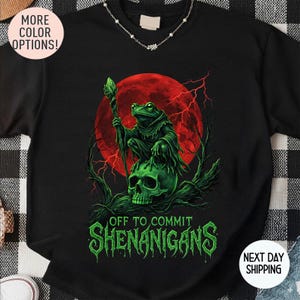 May include: Black sweatshirt featuring a green frog in a robe, holding a staff, atop a green skull, with a red moon backdrop. The text "OFF TO COMMIT SHENANIGANS" is in green. A fun, fantasy-themed design.
