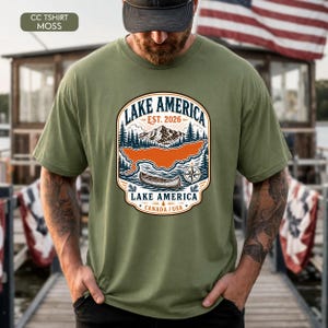 May include: A moss green short-sleeve t-shirt featuring a graphic print of a mountain landscape, a canoe, and a compass. The text reads LAKE AMERICA, EST. 2026, and CANADA / USA. The design includes pine trees and a stylized orange lake shape.