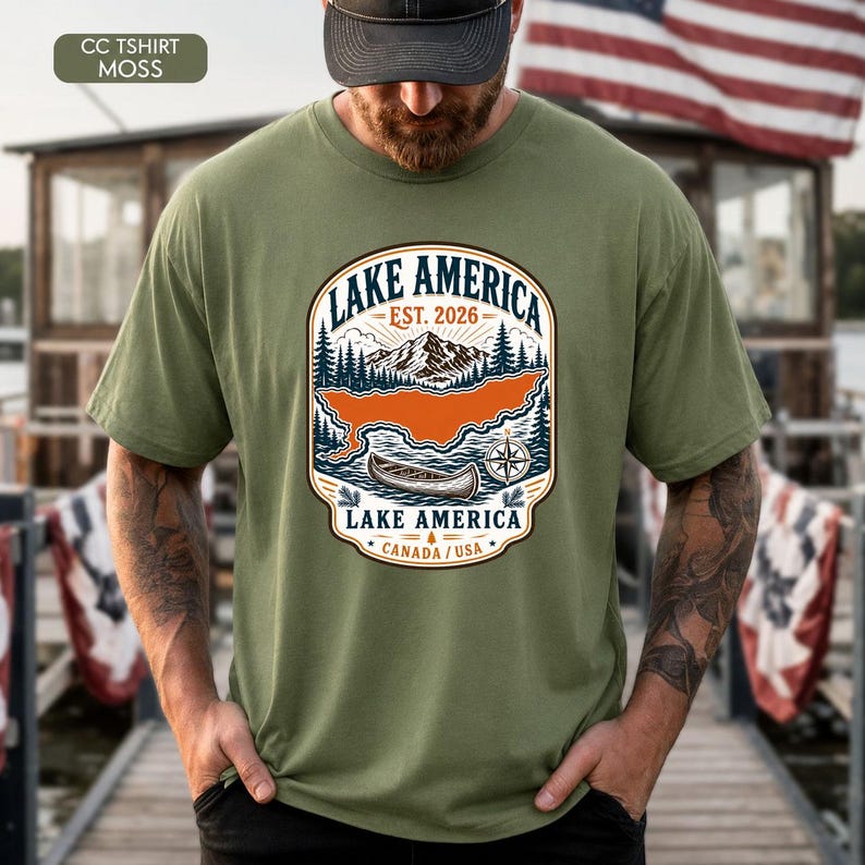 May include: A moss green short-sleeve t-shirt featuring a graphic print of a mountain landscape, a canoe, and a compass. The text reads LAKE AMERICA, EST. 2026, and CANADA / USA. The design includes pine trees and a stylized orange lake shape.