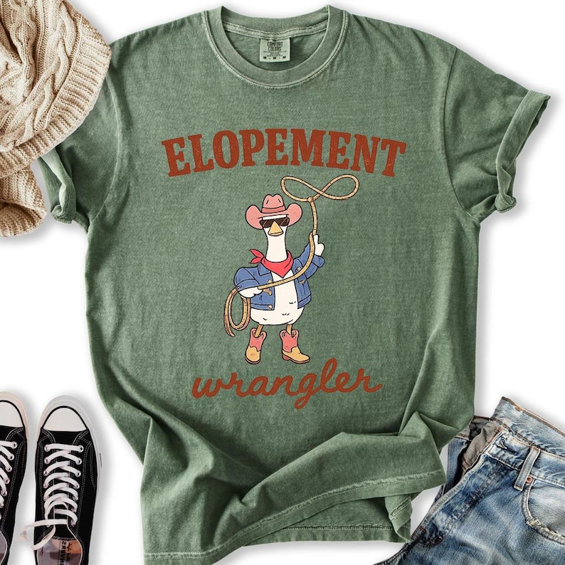 May include: Green t-shirt with the words "ELOPEMENT wrangler" in a rustic font. The shirt features a cartoon goose wearing a cowboy hat, sunglasses, a red bandana, and a denim jacket, holding a lasso.