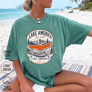 May include: A light green short-sleeve t-shirt featuring a graphic print of a mountain landscape, a canoe, and a compass. The text reads Lake America, Est. 2026, and Canada/USA. The design is enclosed in a decorative border with pine trees.