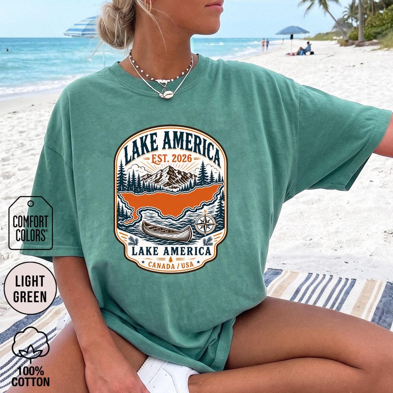 May include: A light green short-sleeve t-shirt featuring a graphic print of a mountain landscape, a canoe, and a compass. The text reads Lake America, Est. 2026, and Canada/USA. The design is enclosed in a decorative border with pine trees.