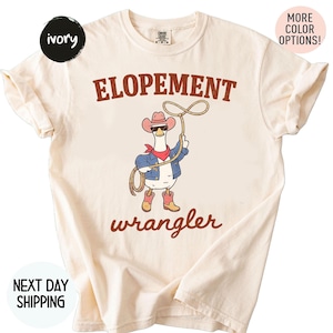 May include: Ivory-colored t-shirt with the words "ELOPEMENT wrangler" in brown. The shirt features a cartoon goose wearing a cowboy hat, sunglasses, a red bandana, and a denim jacket, holding a lasso. The shirt also has the text "MORE COLOR OPTIONS!"