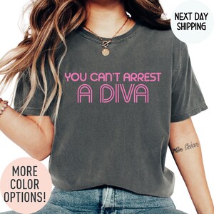 May include: Dark gray t-shirt with the text "YOU CAN'T ARREST A DIVA" in pink. The shirt has a crew neck and short sleeves. The image also includes a necklace and the words "MORE COLOR OPTIONS!"