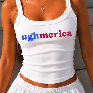 Pu&ograve; includere: Una canotta bianca a coste con la scritta "ughmerica" stampata sul petto. La parola "ugh" &egrave; blu, mentre "merica" &egrave; di una tonalit&agrave; rosa rossastra. Il capo presenta uno scollo rotondo e spalline larghe, offrendo una vestibilit&agrave; aderente.