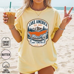 May include: A butter yellow short-sleeve t-shirt featuring a graphic print of a mountain landscape, a canoe, and a compass. The text reads LAKE AMERICA, EST. 2026, and CANADA / USA. The design includes dark blue, orange, and white ink colours.
