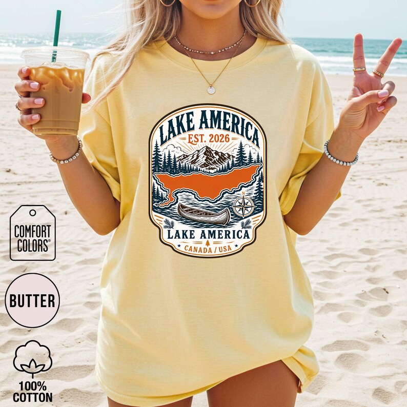 May include: A butter yellow short-sleeve t-shirt featuring a graphic print of a mountain landscape, a canoe, and a compass. The text reads LAKE AMERICA, EST. 2026, and CANADA / USA. The design includes dark blue, orange, and white ink colours.