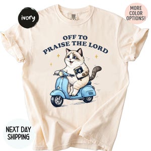 May include: Ivory-colored t-shirt featuring a cartoon cat riding a light blue scooter, holding a book, and the text "OFF TO PRAISE THE LORD". The shirt has rolled-up sleeves and a round neckline.
