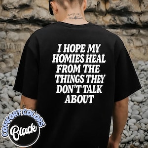 May include: A black short-sleeved t-shirt featuring white text on the back that reads, I HOPE MY HOMIES HEAL FROM THE THINGS THEY DON'T TALK ABOUT. The shirt has a crew neckline and a standard fit, displayed from a rear-facing perspective.