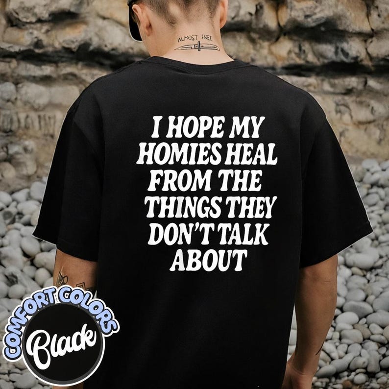 May include: A black short-sleeved t-shirt featuring white text on the back that reads, I HOPE MY HOMIES HEAL FROM THE THINGS THEY DON'T TALK ABOUT. The shirt has a crew neckline and a standard fit, displayed from a rear-facing perspective.