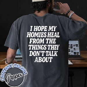 May include: A charcoal gray short-sleeve t-shirt featuring white text on the back that reads, I HOPE MY HOMIES HEAL FROM THE THINGS THEY DON'T TALK ABOUT. The shirt has a relaxed fit and a solid color design with a circular logo in the corner.